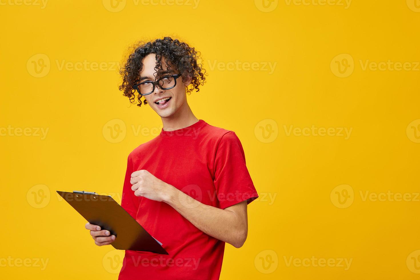 alegre contento miope joven estudiante hombre en rojo camiseta gracioso gafas sostiene tableta carpeta con estudiar notas posando aislado en terminado amarillo estudio antecedentes. gratis sitio para anuncio. educación Universidad concepto foto