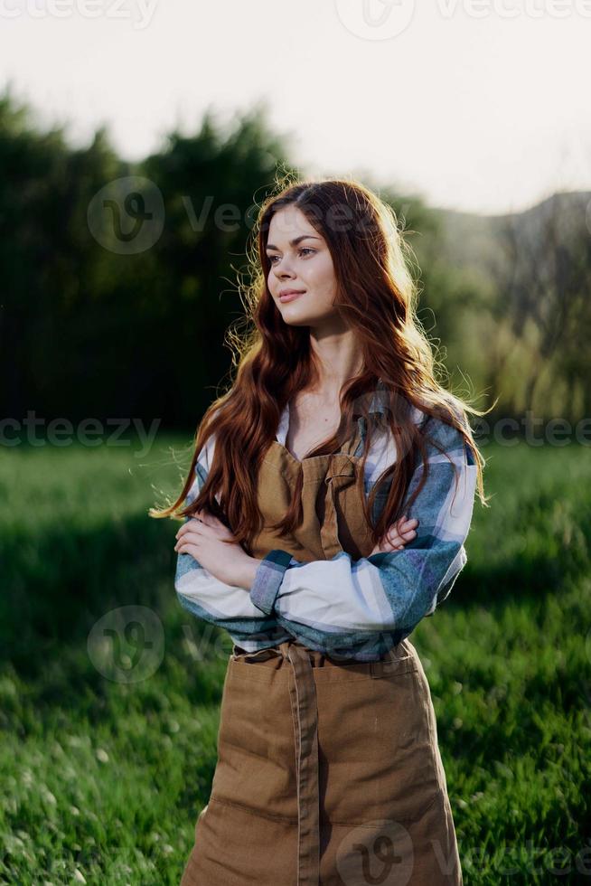 A farmer woman in her work clothes, plaid shirt and apron, stands in the field on the green grass and smiles in the setting sun photo