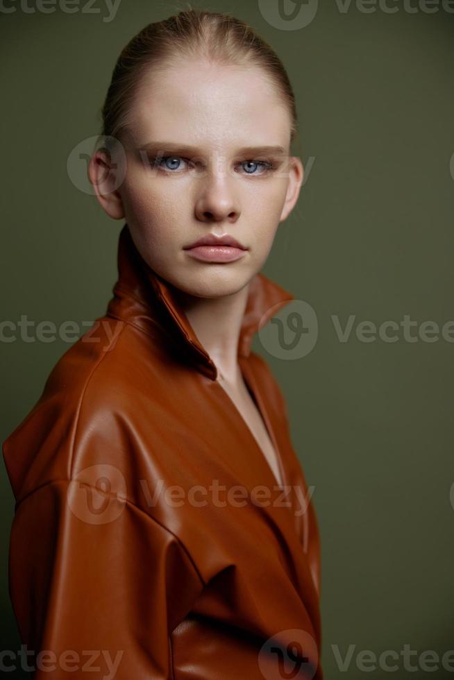 Stylish serious young blonde girl with blue eyes with evening makeup in a leather dress looks at the camera posing isolated. Studio portrait on a green background. Fashion offer photo