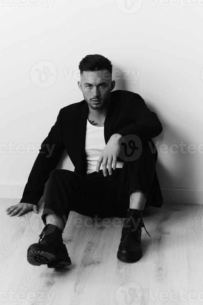 Modelling snapshots. Serious self-confident attractive handsome man in black jacket sitting on floor posing in white studio background. Black and White concept. Copy space photo