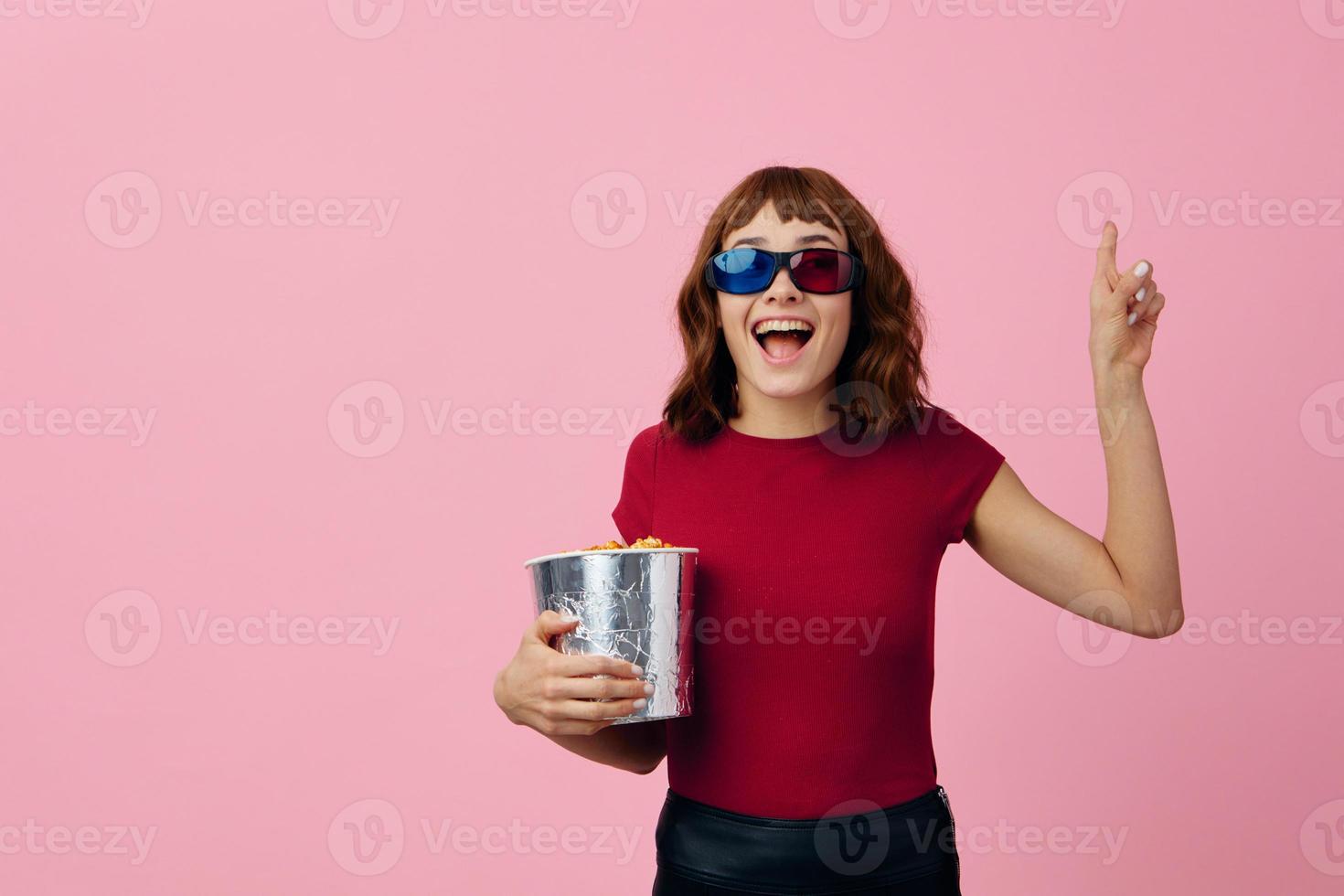 lleno de alegría contento linda pelirrojo dama en rojo camiseta 3d lentes con palomitas de maiz reloj película posando aislado en terminado rosado estudio antecedentes. Copiar espacio bandera. Moda cine concepto. entretenimiento oferta foto