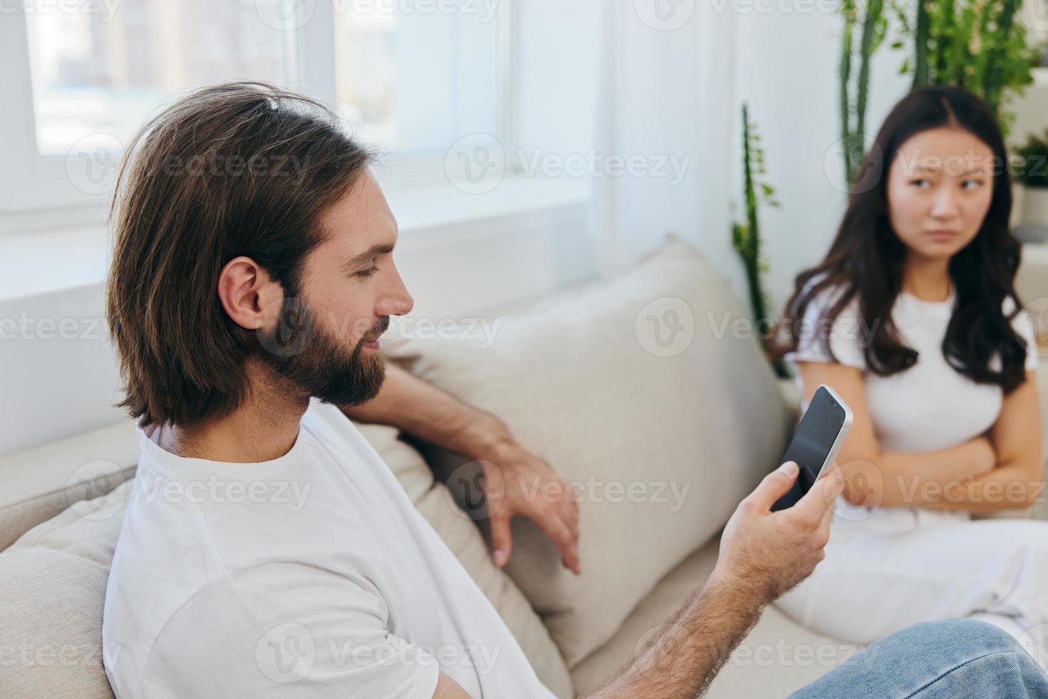 A man looks at his phone screen and smiles during an argument with his girlfriend. The angry and hurt woman looks in his direction and is sad. Family discord at home, phone addiction photo