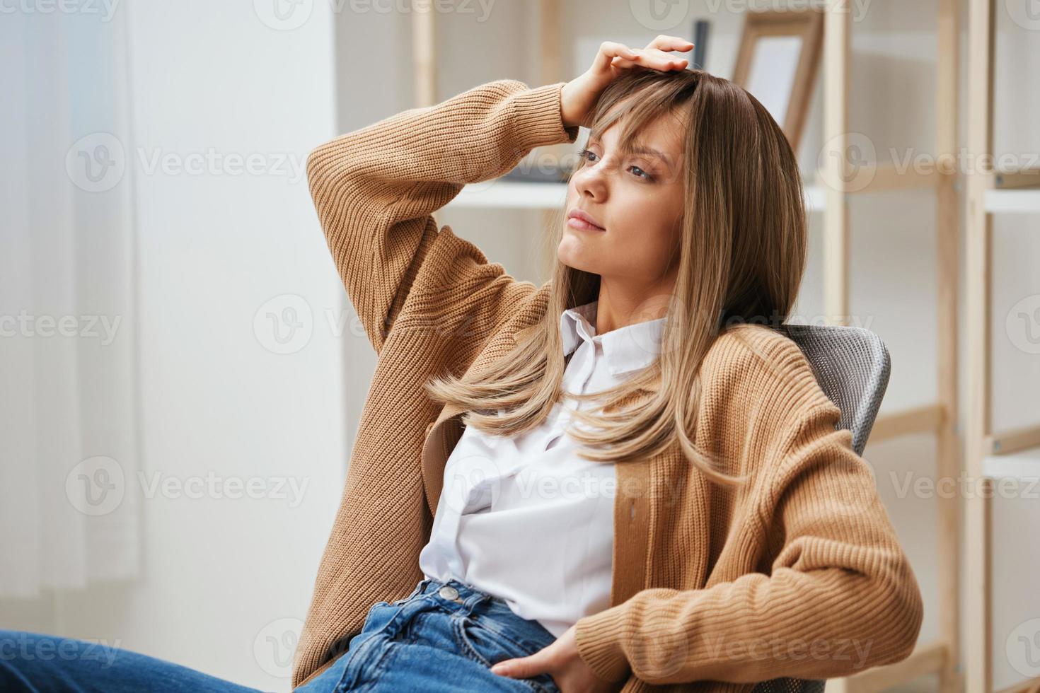 Thoughtful pensive young blonde lady in warm sweater dreams about future holidays sitting in armchair at home interior. Pause from work, take a break, reflections in free time concept. Copy space photo