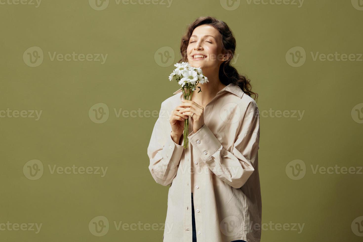 Smiling cute curly beautiful female in linen casual shirt hold chamomiles flowers near face enjoying smell posing isolated on over olive green pastel background. Nature is beauty concept. Copy space photo