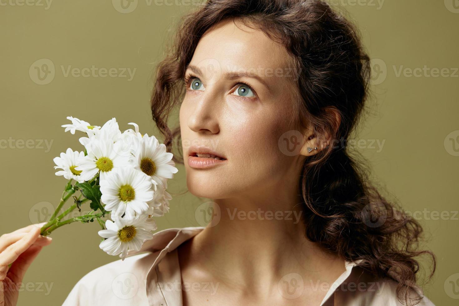 Close up portrait of adorable curly female in linen casual shirt hold chamomiles flowers near face looks up posing isolated on over olive green pastel background. Nature is beauty concept. Copy space photo