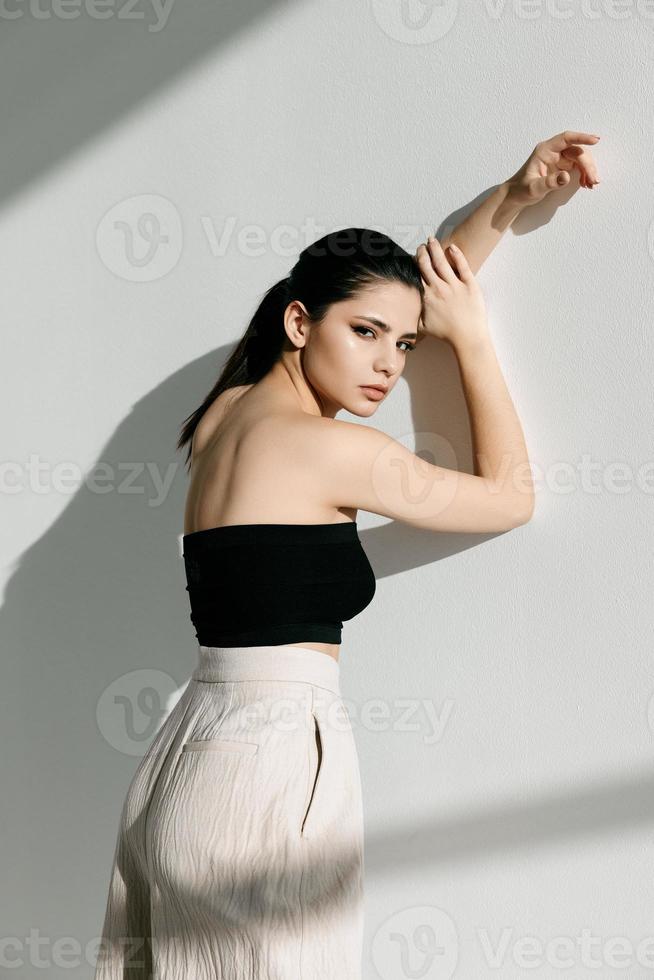 stylish woman in white pants and a short T-shirt leaned against the wall and a bright light photo