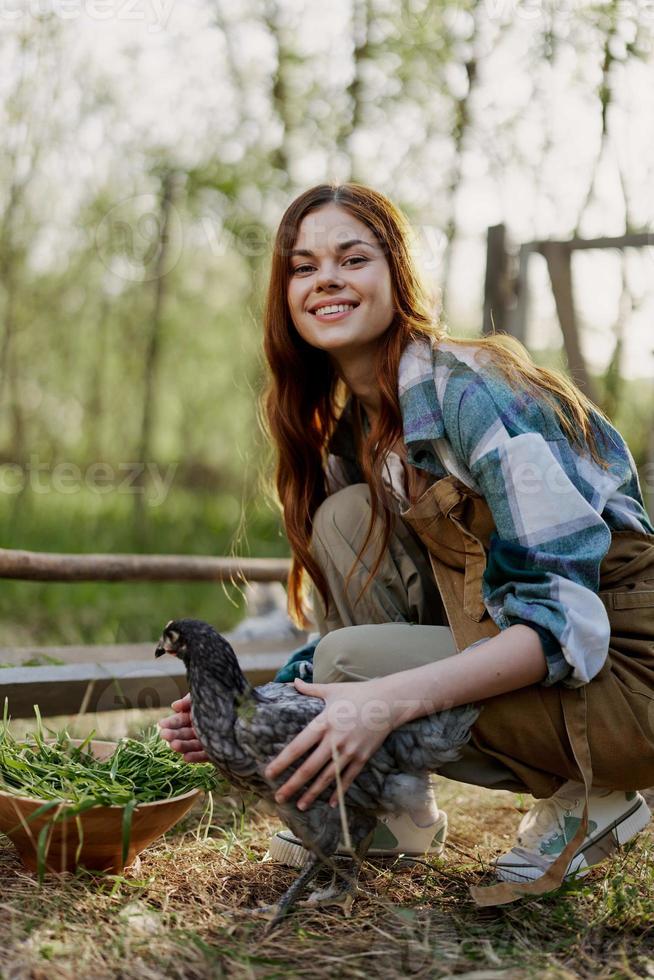 Woman smiles looking at the chicken she holds near the feeder in her hands on the farm, farm labor for raising healthy birds and feeding them organic food in nature photo