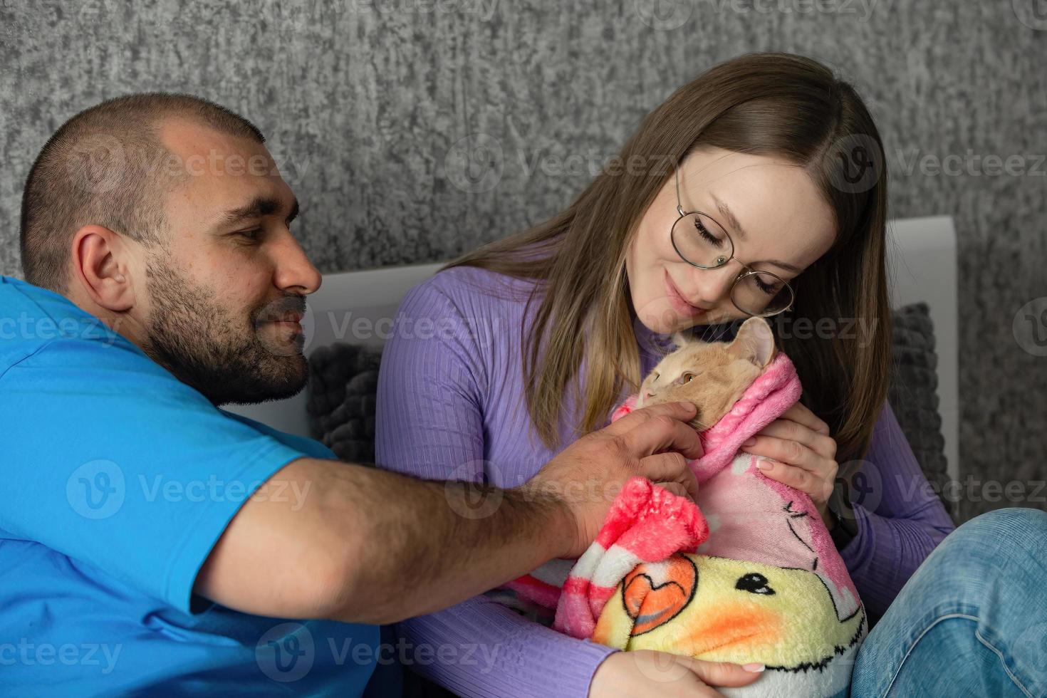 un joven Pareja en el cama abrazos su gato, envuelto en un bebé frazada. el de pareja consciente rechazo a tener niños, tratando mascotas me gusta niños. foto