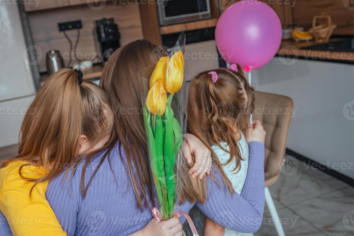 Daughters congratulate their mom on Mother's Day, a card with a heart, flowers and a balloon at home in the kitchen, they all hug. Children surprise their mother for the holiday. photo