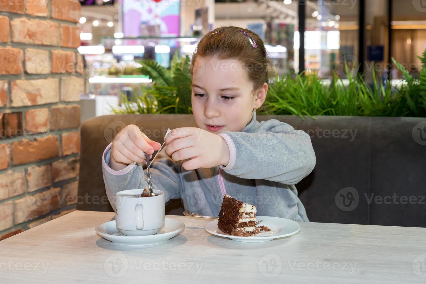 pequeño niña en un café a un mesa con un postre toma fuera un té bolso desde un taza foto