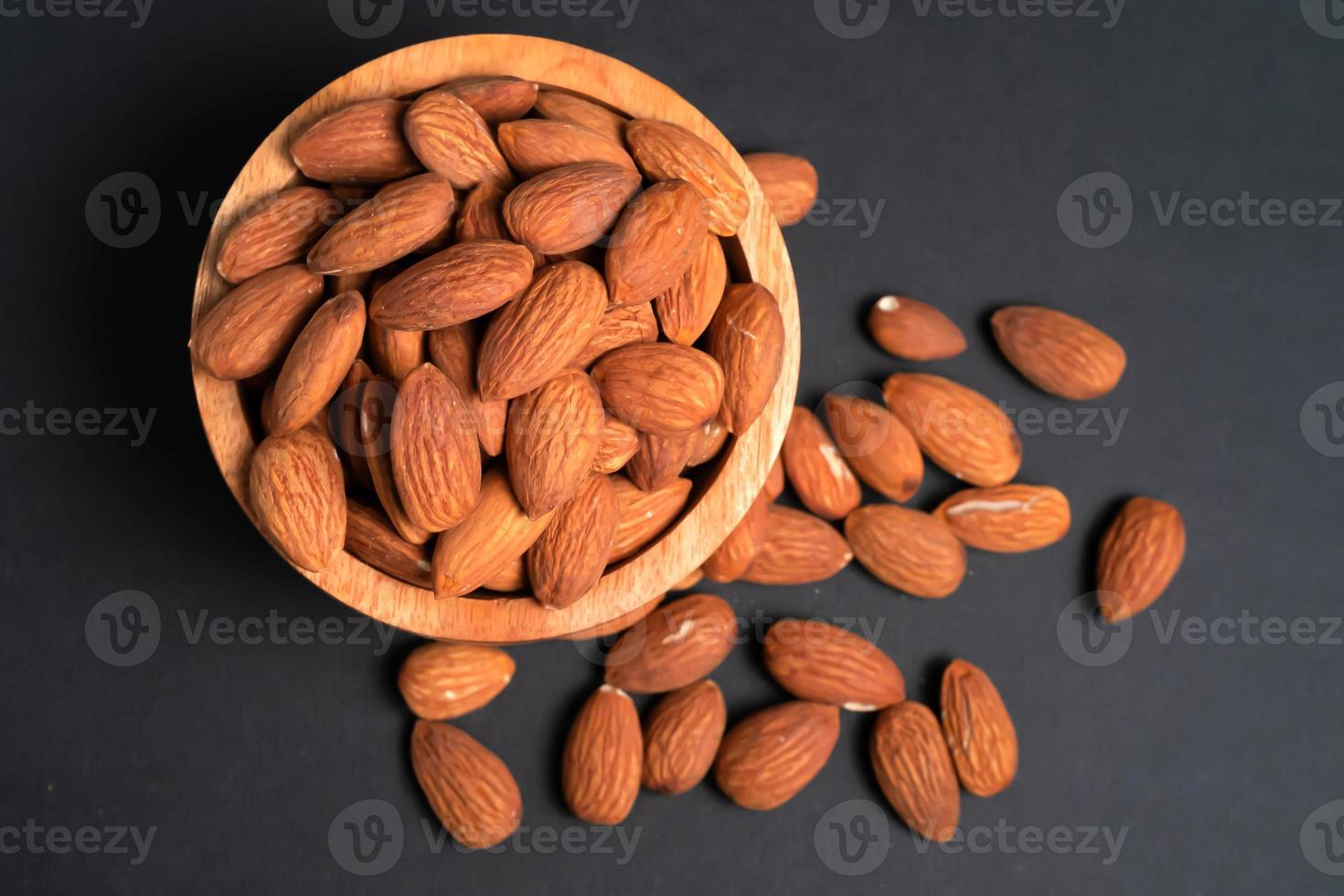 Peeled almonds in a wooden bowl and some scattered nuts on black background, top view. photo