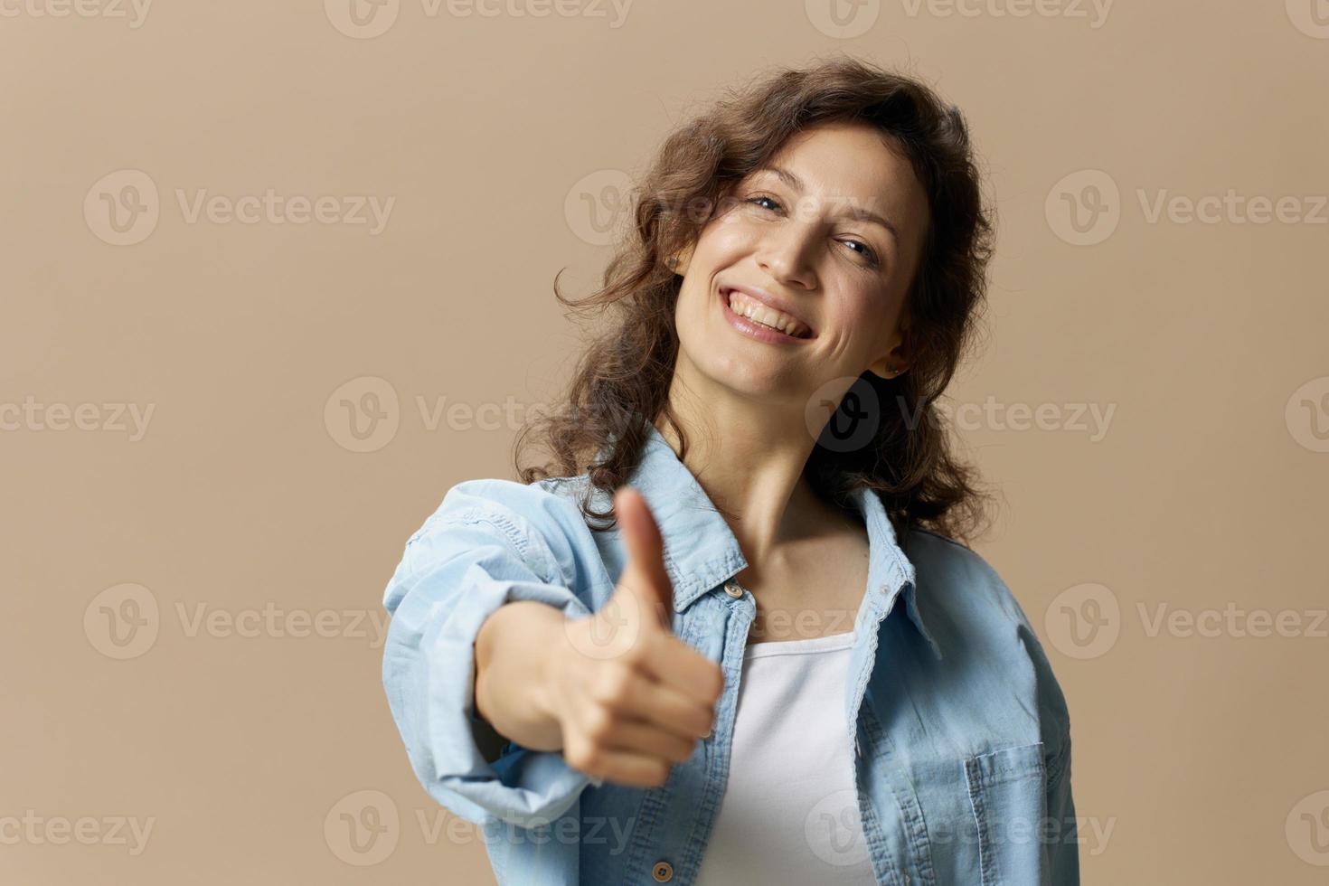 Happy smiling curly beautiful female in jeans casual shirt show thumb up gesture Like at camera posing isolated on over beige pastel background. People Lifestyle emotions concept. Copy space photo