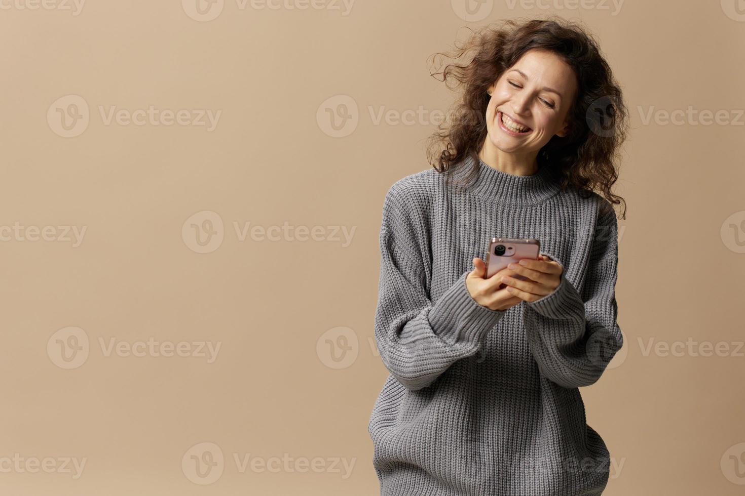 Lovely happy curly beautiful female in gray casual sweater chatting with boyfriend posing isolated on over beige pastel background. Social media, network, distance communication concept. Copy space photo