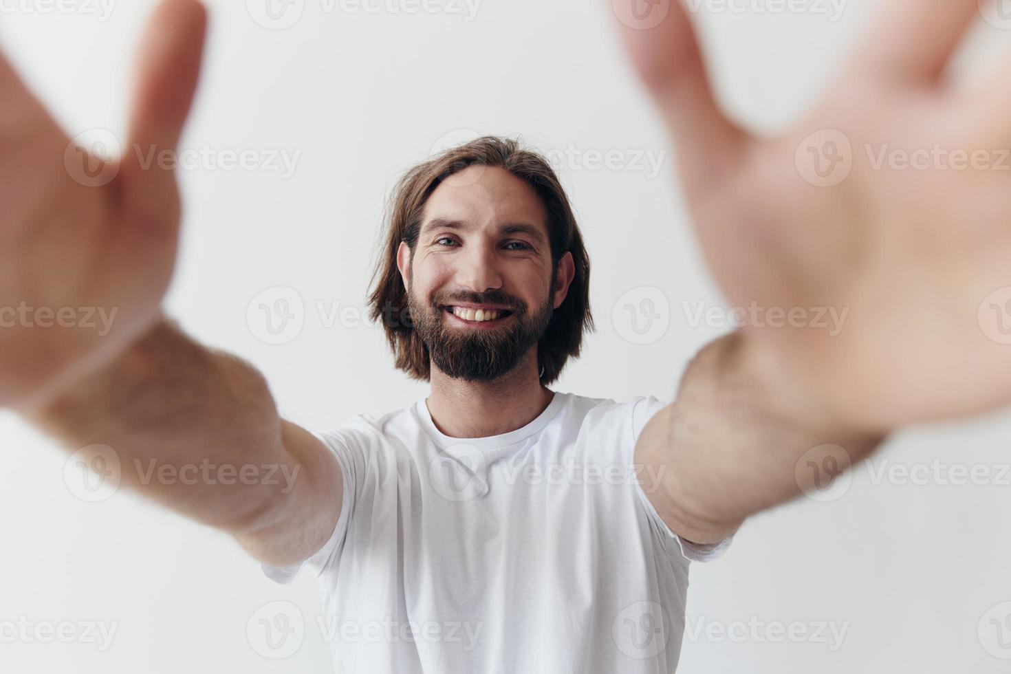 Happy adult man with a beard smiles and pulls the cook into the camera listening to music in headphones in a distressed t-shirt on a white isolated background photo