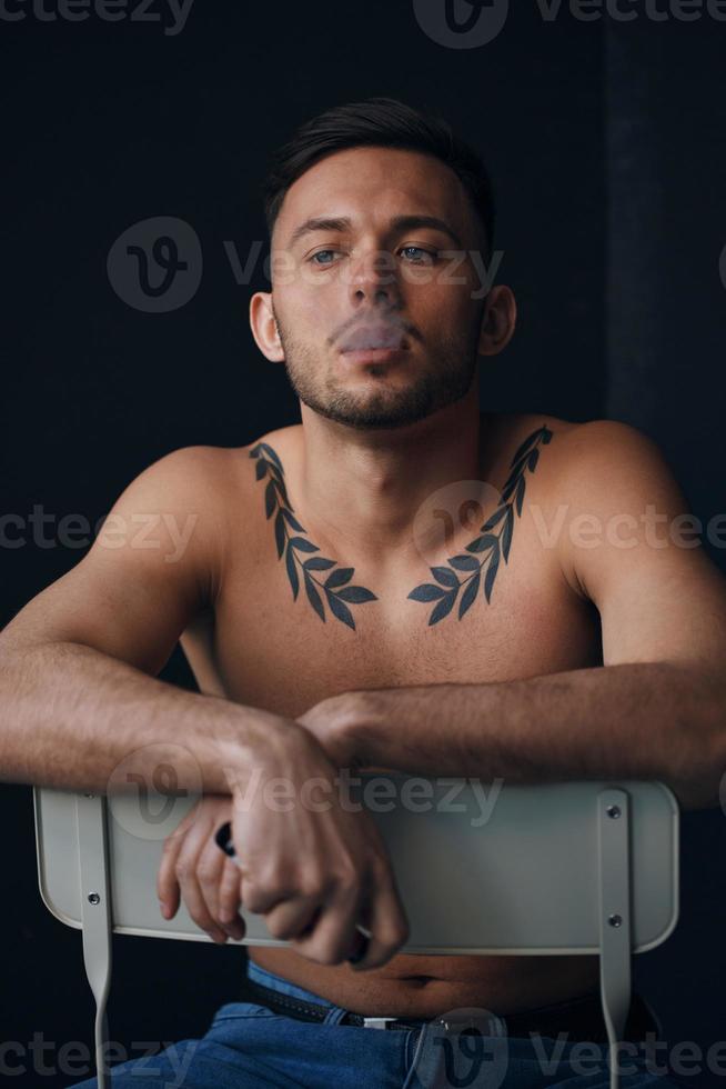 Modelling snapshots. Pensive serious tanned attractive handsome naked man sit on chair smoking looks aside posing isolated in black studio background. Fashion offer. Copy space for ad. Closeup photo