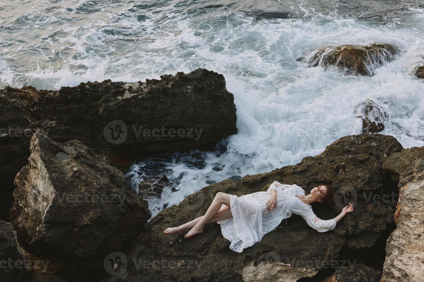 atractivo mujer con largo pelo en un blanco vestir acostado en un Roca en un blanco vestir inalterado foto