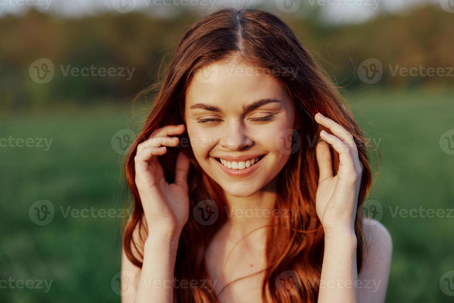 A beautiful woman with long, wavy red hair smiles for the camera in nature illuminated by sunset sunlight. The concept of beauty and harmony with nature photo