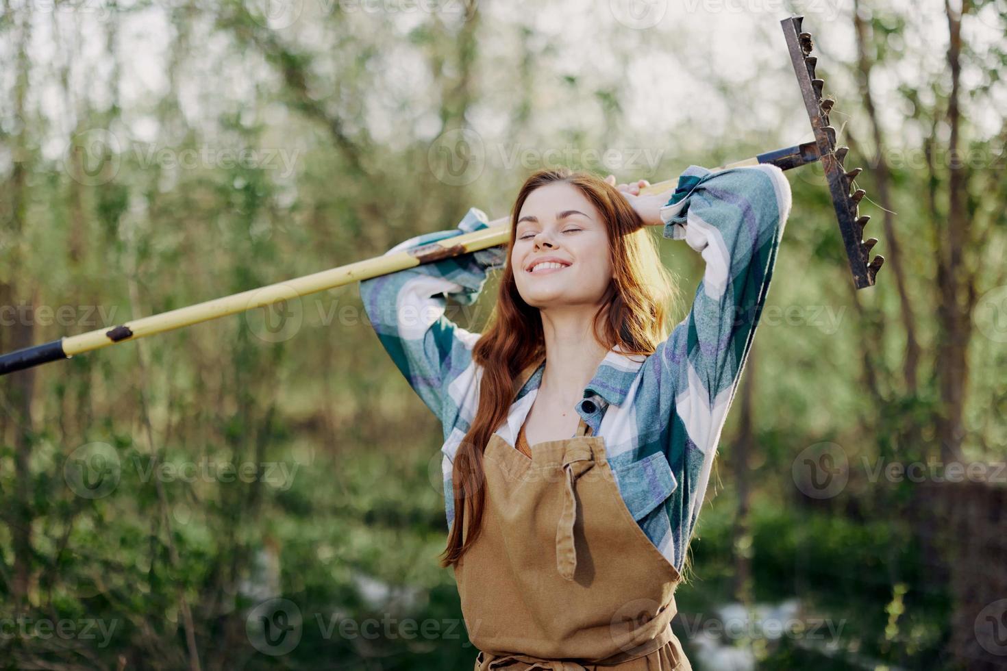 A woman smiling beautifully and looking at the camera, a farmer in work clothes and an apron working outdoors in nature and holding a rake to gather grass and forage for the animals in the garden photo