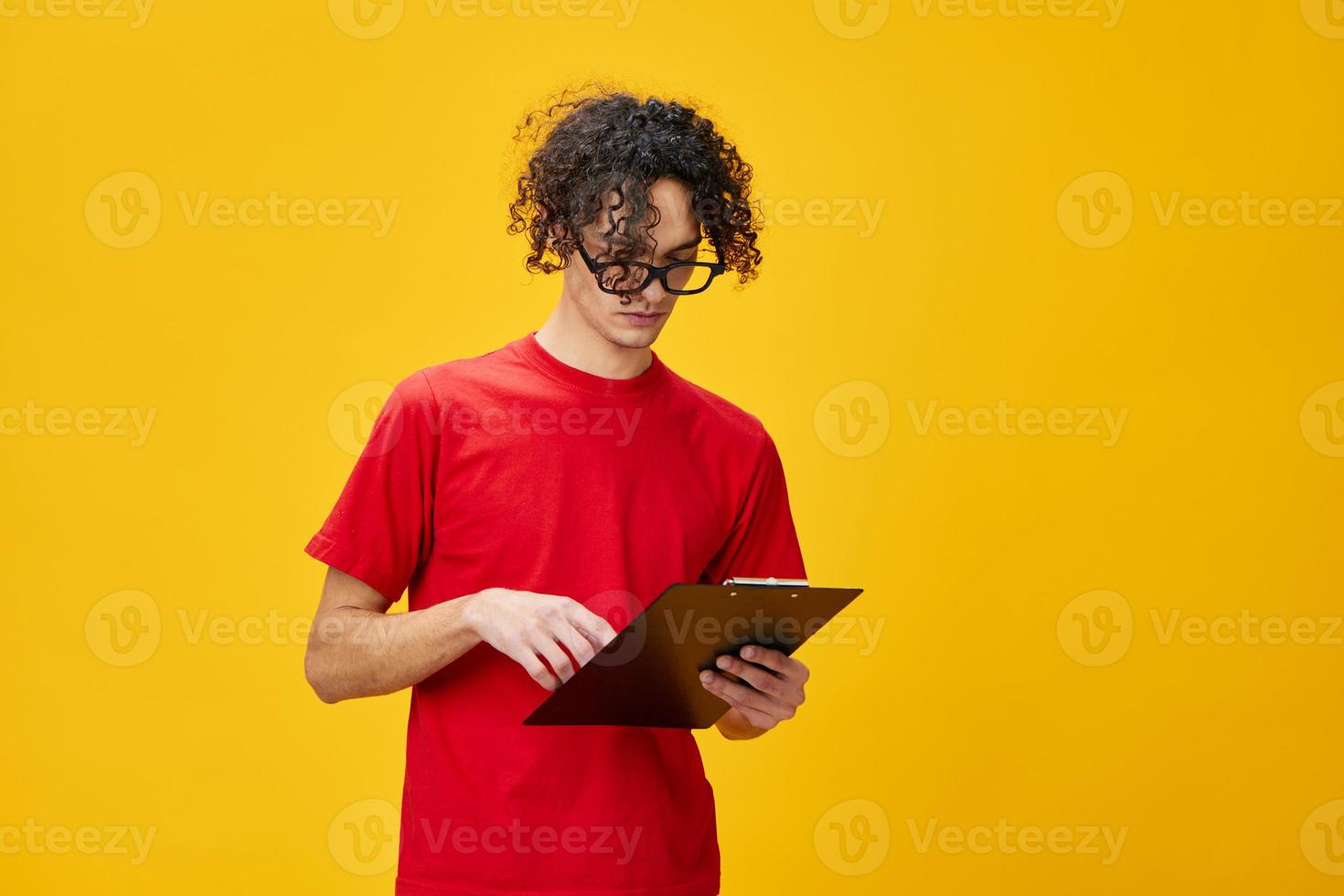 confuso miope joven estudiante hombre en rojo camiseta gracioso gafas sostiene tableta carpeta con estudiar notas posando aislado en terminado amarillo estudio antecedentes. gratis sitio para anuncio. educación Universidad concepto foto