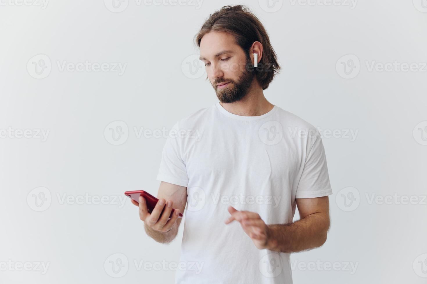 A man with a hipster beard in a white T-shirt with a phone and wireless headphones smiling listening to music and an audiobook online against a white wall photo