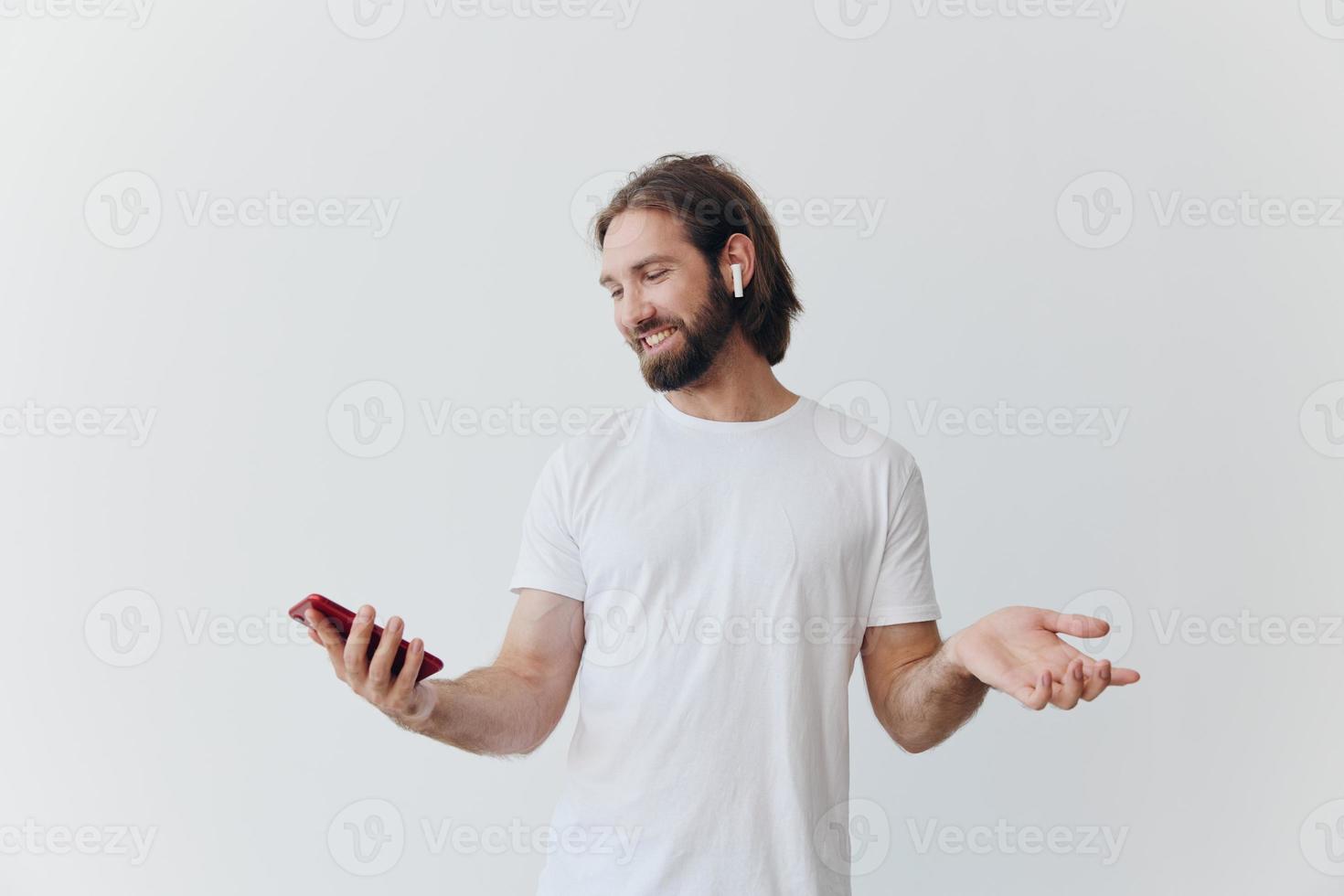 A man with a hipster beard in a white T-shirt with a phone and wireless headphones smiling listening to music and an audiobook online against a white wall photo