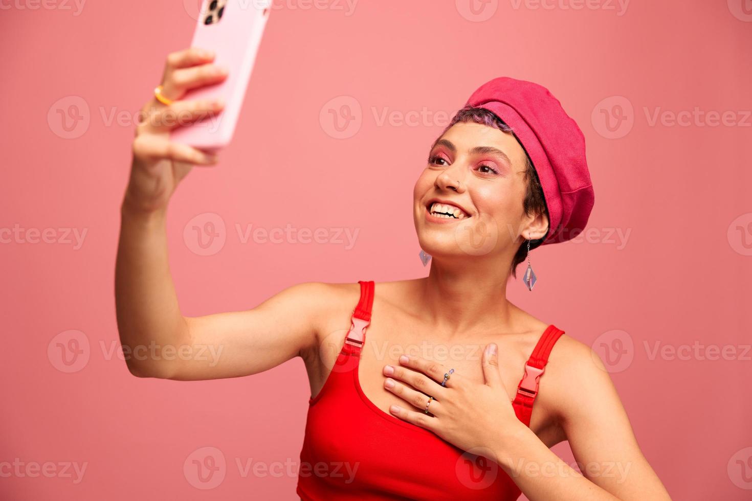 A young woman blogger with colored pink hair and a short haircut takes a picture of herself on the phone and broadcasts a smile in stylish clothes and a hat on a pink background monochrome style photo