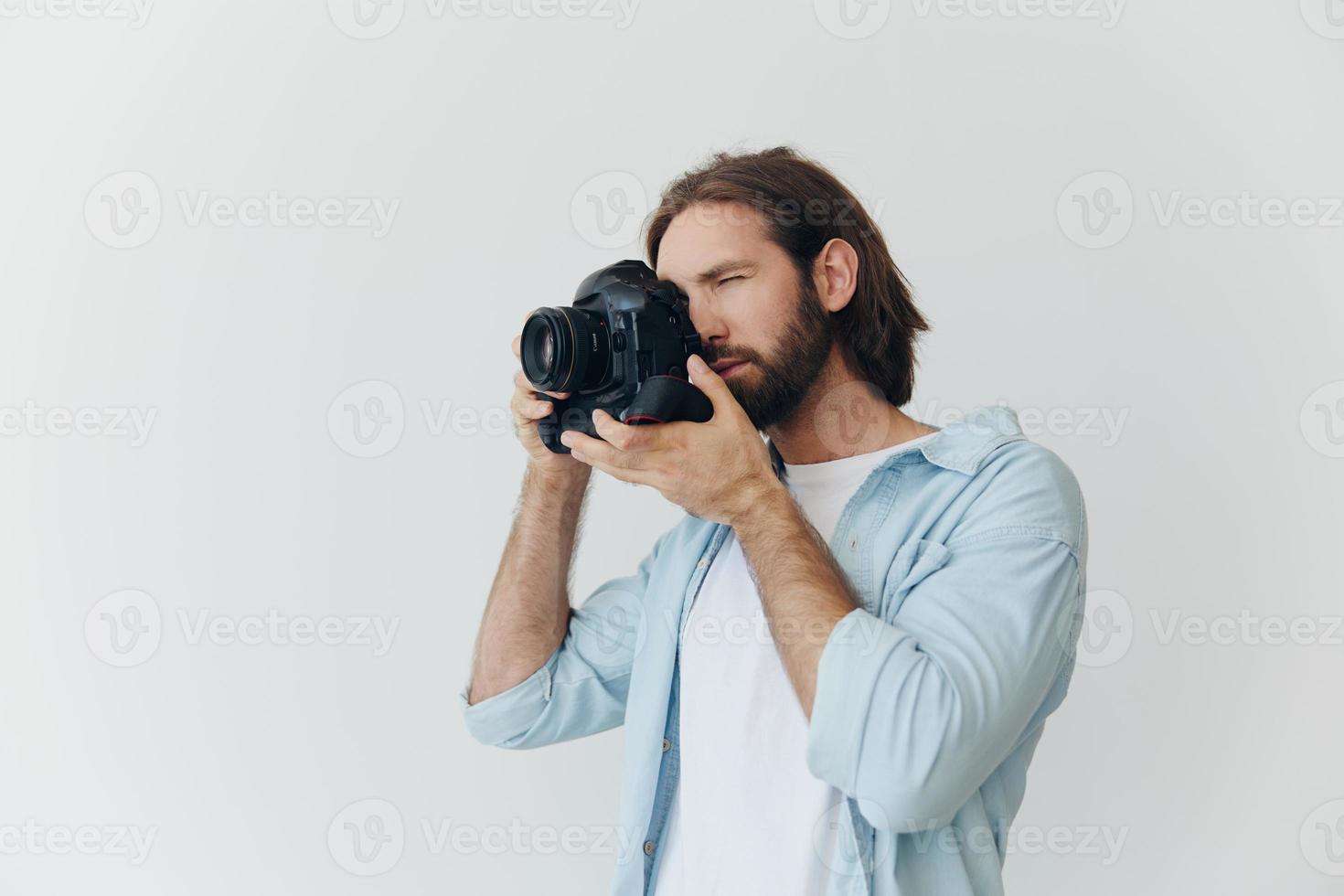 A male hipster photographer in a studio against a white background looks through the camera viewfinder and shoots shots with natural light from the window. Lifestyle work as a freelance photographer photo