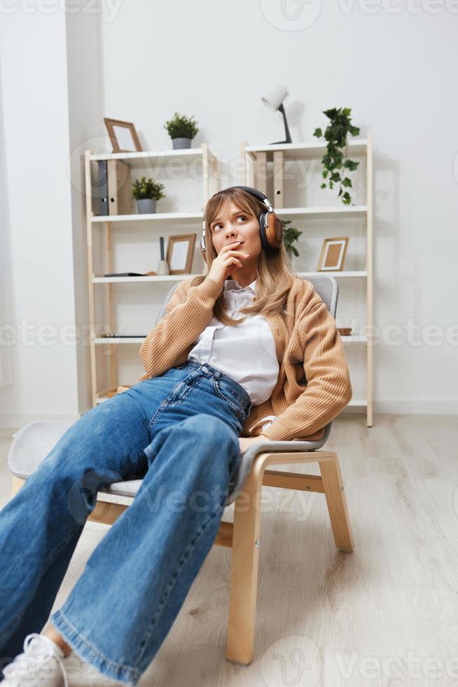 Dreaming happy young blonde lady in warm sweater in headphones looks aside listen fav songs sitting in armchair at modern home interior. Music time Relaxing Cool playlist Concept. Copy space photo