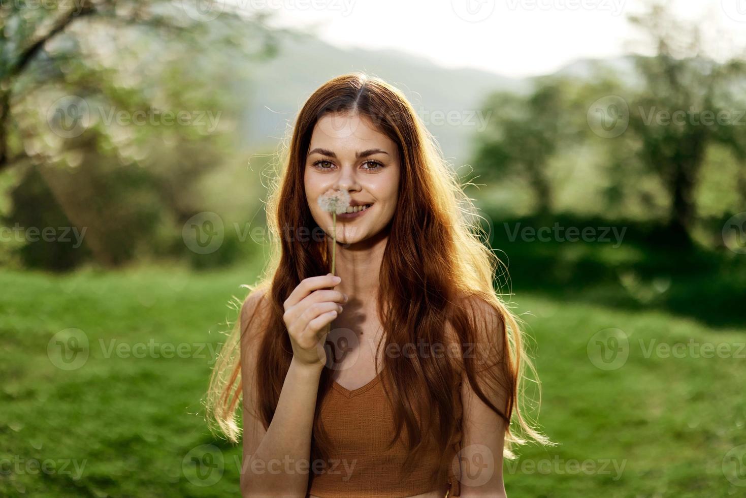 Portrait of a young woman with a dandelion flower in her hand blowing on it and smiling against the green summer grass in the rays of the setting sun in nature photo