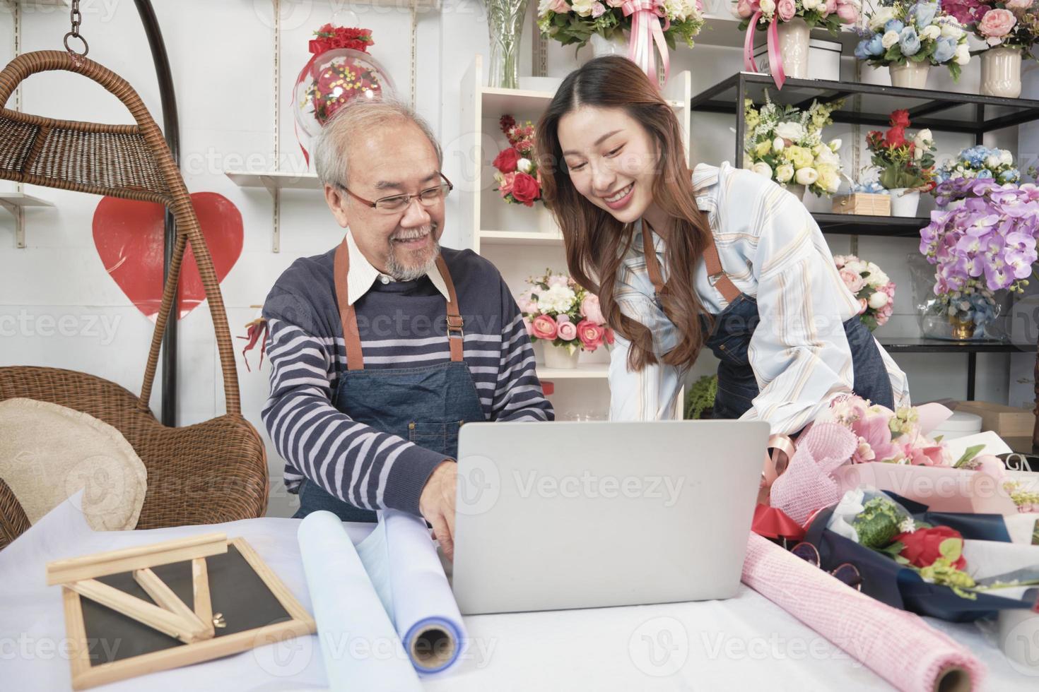 Asian elder male florist owner discussing with young beautiful female employee who shopkeeper about website arrangement for business service, happy work in colorful flower shop store, e-commerce SME. photo