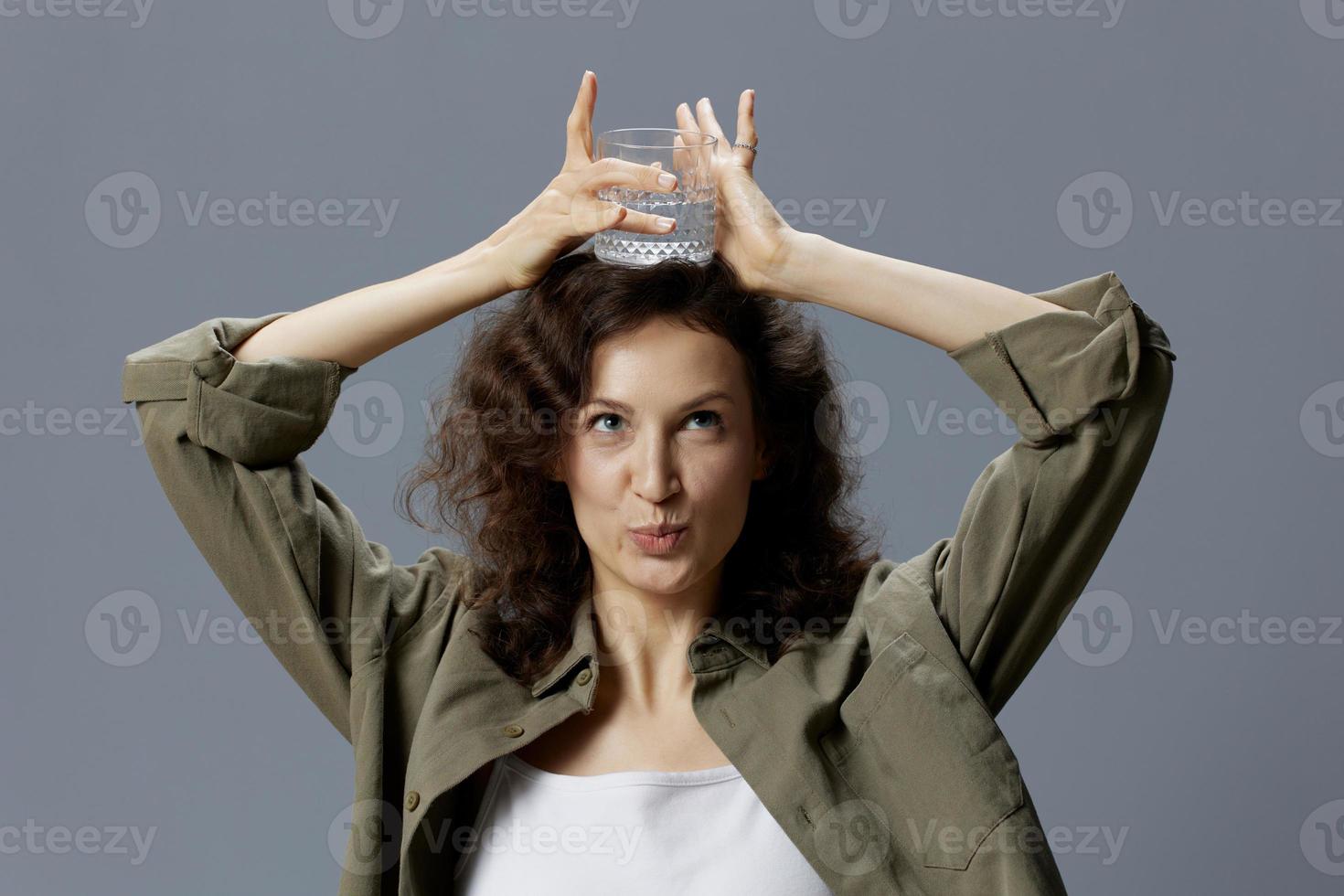 Funny pretty curly beautiful woman in casual khaki green shirt hold glass of water overhead posing isolated on over gray blue background. Healthy lifestyle. Water is Life concept. Copy space photo