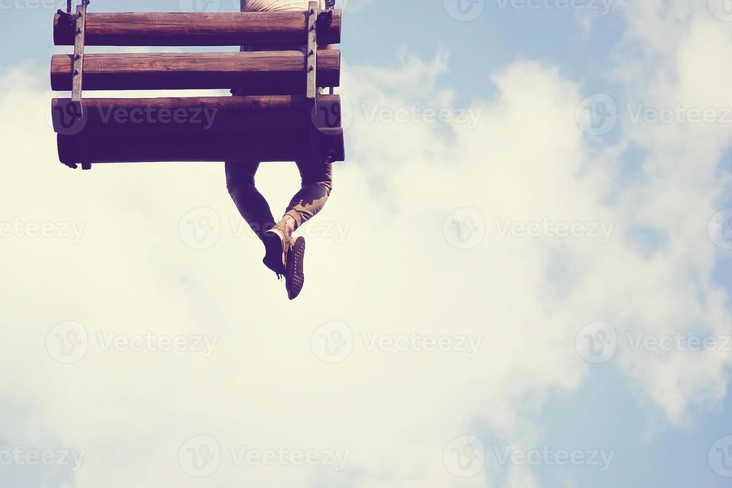 One young man sitting on wooden swing in the sky with his legs crossed. Back view, copy space. photo