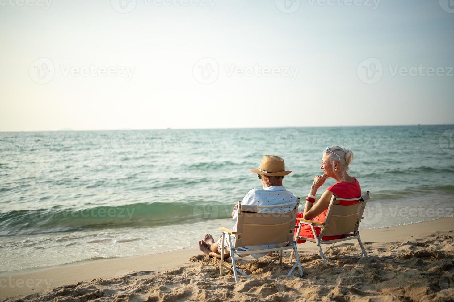 A couple of elderly sitting in chairs at the beach watching the sun and the sea on their summer vacation and they smile and enjoy their vacation. photo