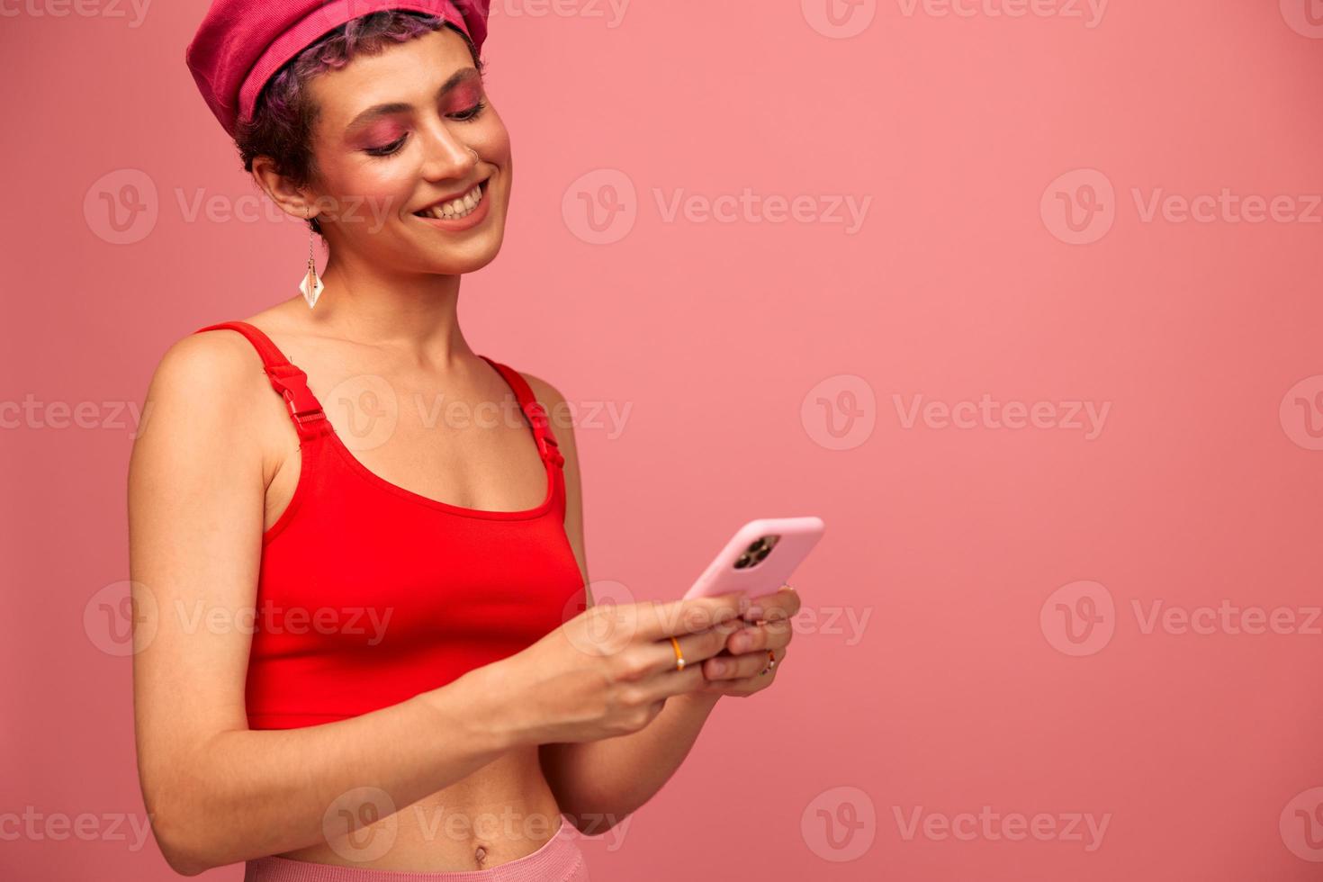 joven mujer blogger con de colores rosado pelo y corto Corte de pelo voltear mediante el pantalla de el teléfono y mecanografía un mensaje con un sonrisa en elegante ropa en un rosado antecedentes foto
