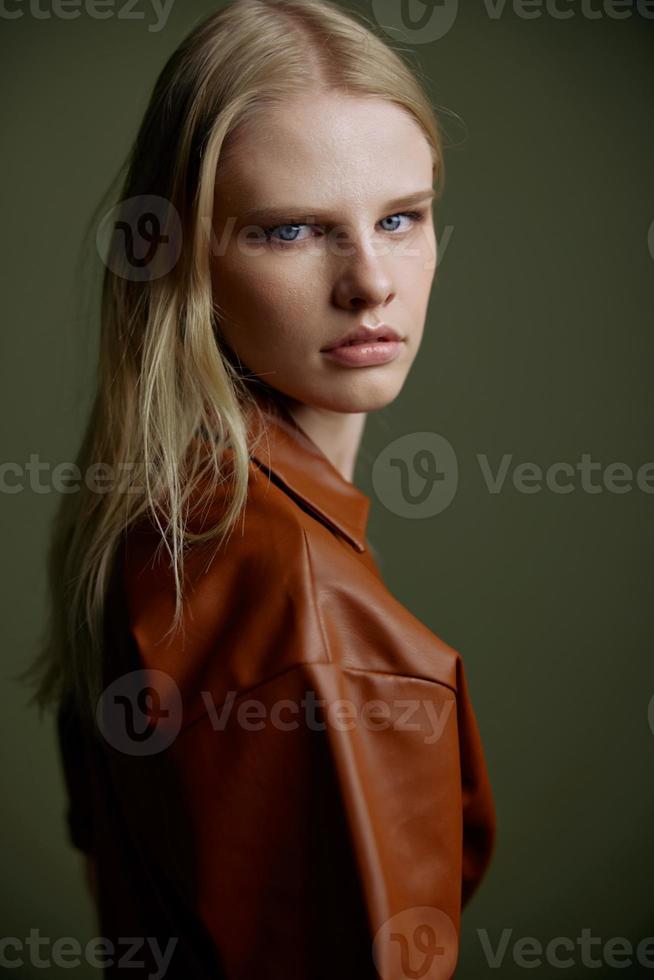 Closeup studio portrait of blonde with expressive glance blue eyes showy makeup looking over her shoulder at camera. Beautiful young woman in trendy leather brown dress posing isolated on green wall photo