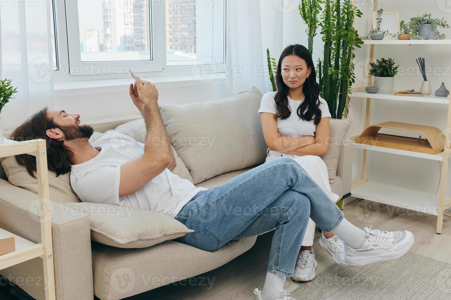 A man looks at his phone screen and smiles during an argument with his girlfriend. The angry and hurt woman looks in his direction and is sad. Family discord at home, phone addiction photo