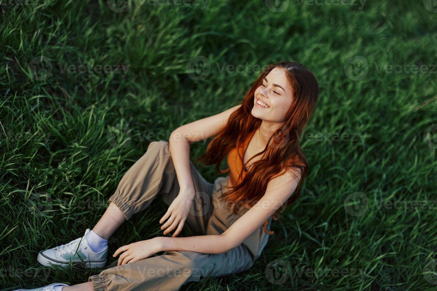 contento mujer sonriente hermosamente y mirando arriba a el cámara sentado en Fresco verde césped en el verano Brillo Solar foto