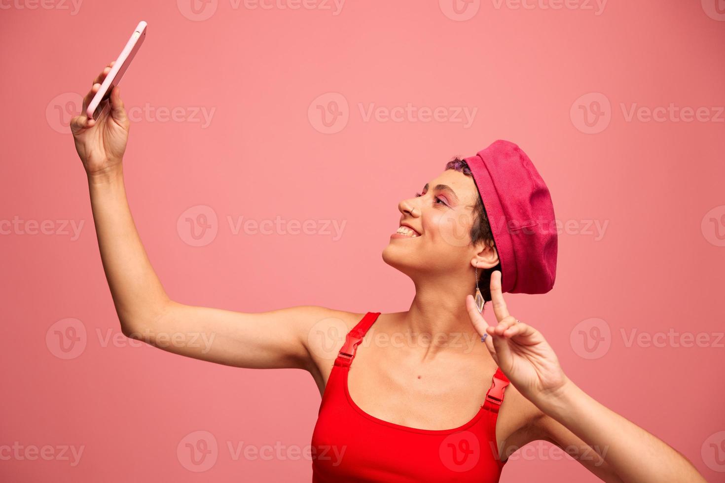 A young woman blogger with colored pink hair and a short haircut takes a picture of herself on the phone and broadcasts a smile in stylish clothes and a hat on a pink background monochrome style photo