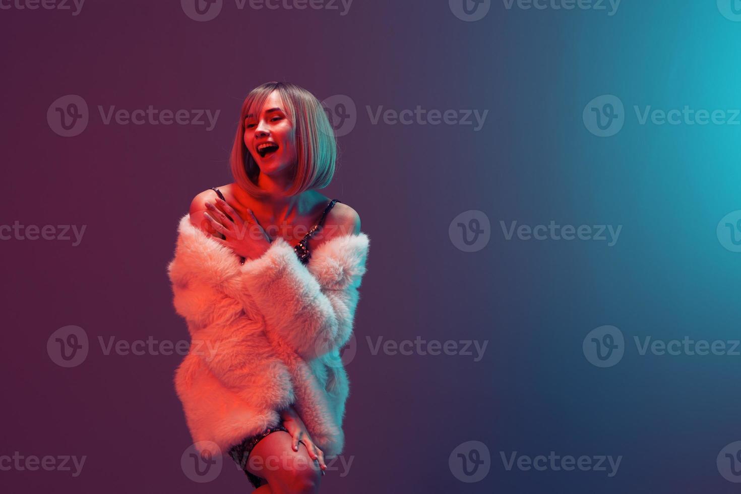Excited happy adorable blonde woman in fluffy fur coat sparkly dress touch herself posing isolated in blue turquoise red color light studio background. Neon party Fashion concept. Copy space Banner photo
