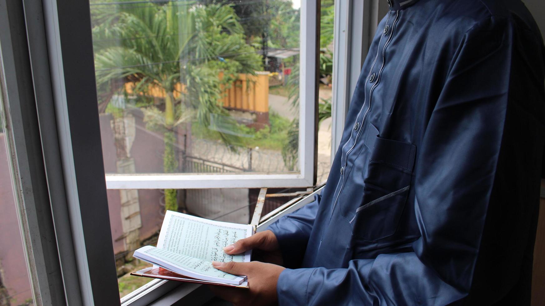 A young man in a muslin shirt is reading the Koran by a glass window. photo