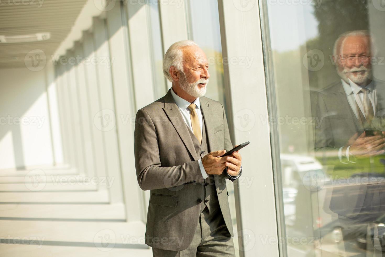 A senior business man stands in an office hallway, focused on his mobile phone photo