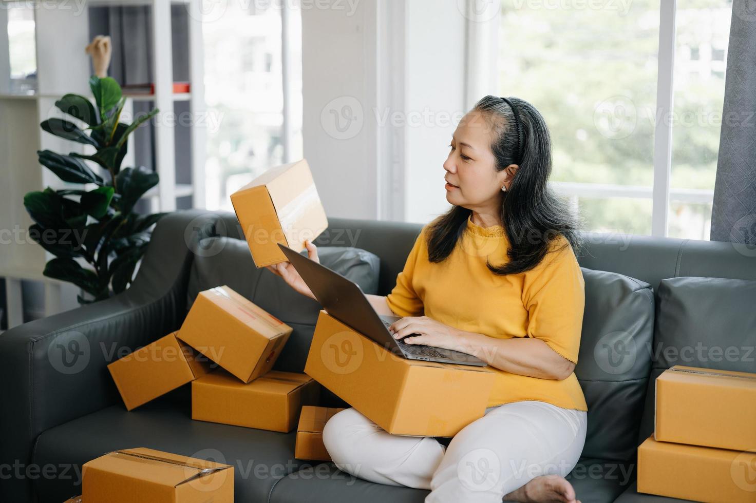 mayor negocio propietario mujer preparar paquete o empaquetar caja y en pie cheque en línea pedidos para entregar a cliente en tableta, ordenador portátil compras en línea concepto. foto