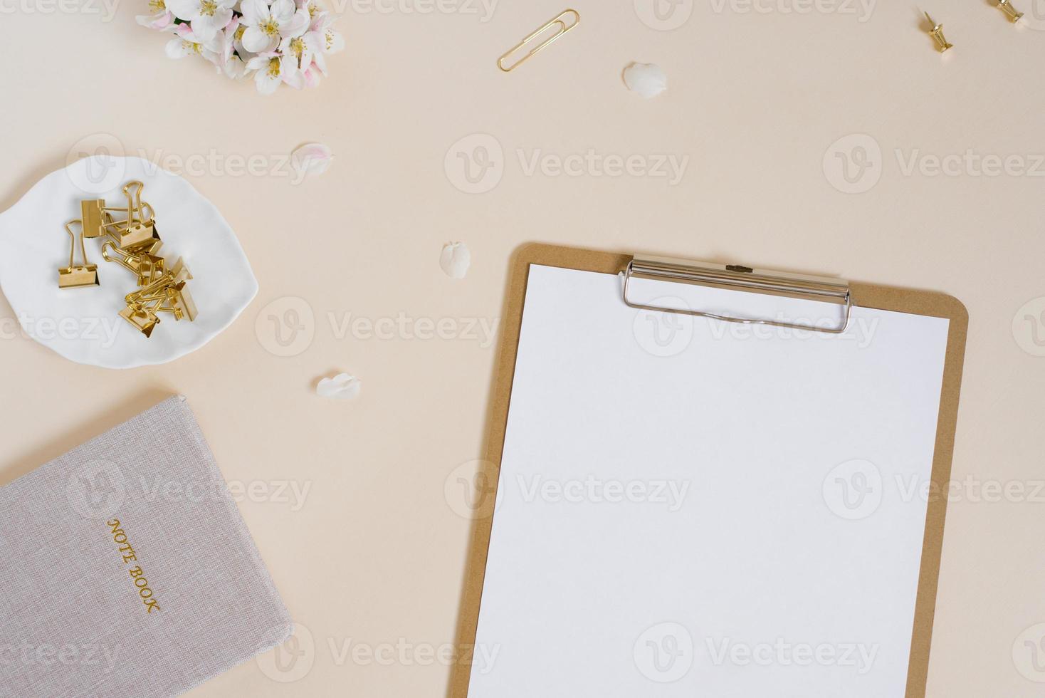 Stylized feminine or blogger desk, office desk. Workplace, white flowers of an apple tree, notebook, pen, paper clips and a tablet with a blank white sheet of paper on a beige background photo