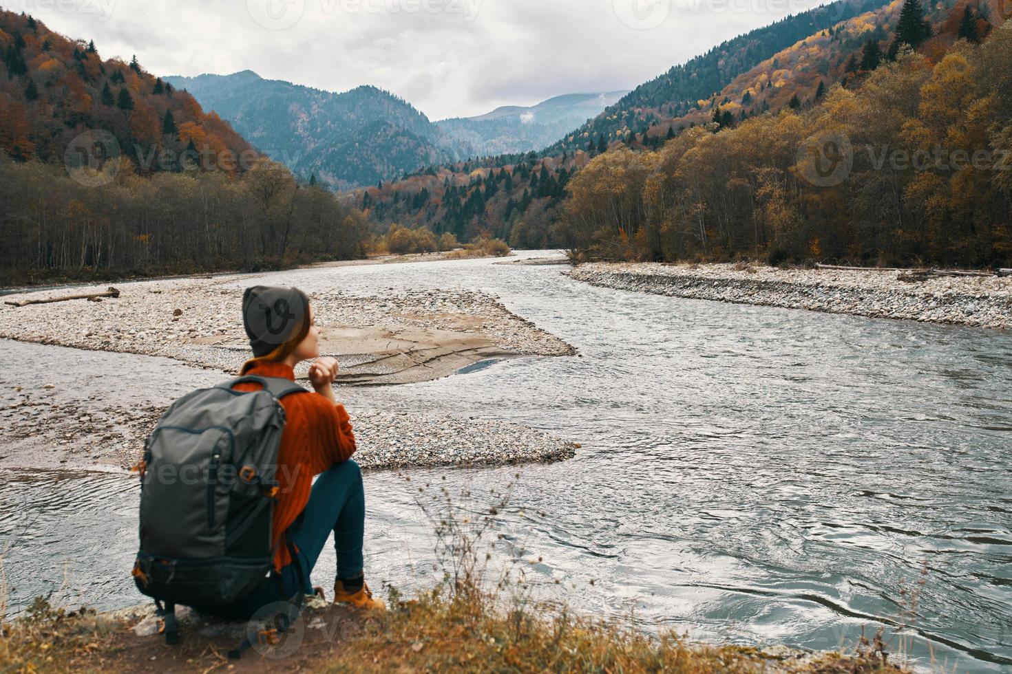 woman traveler with a backpack near the river on the grass and mountains in the distance photo