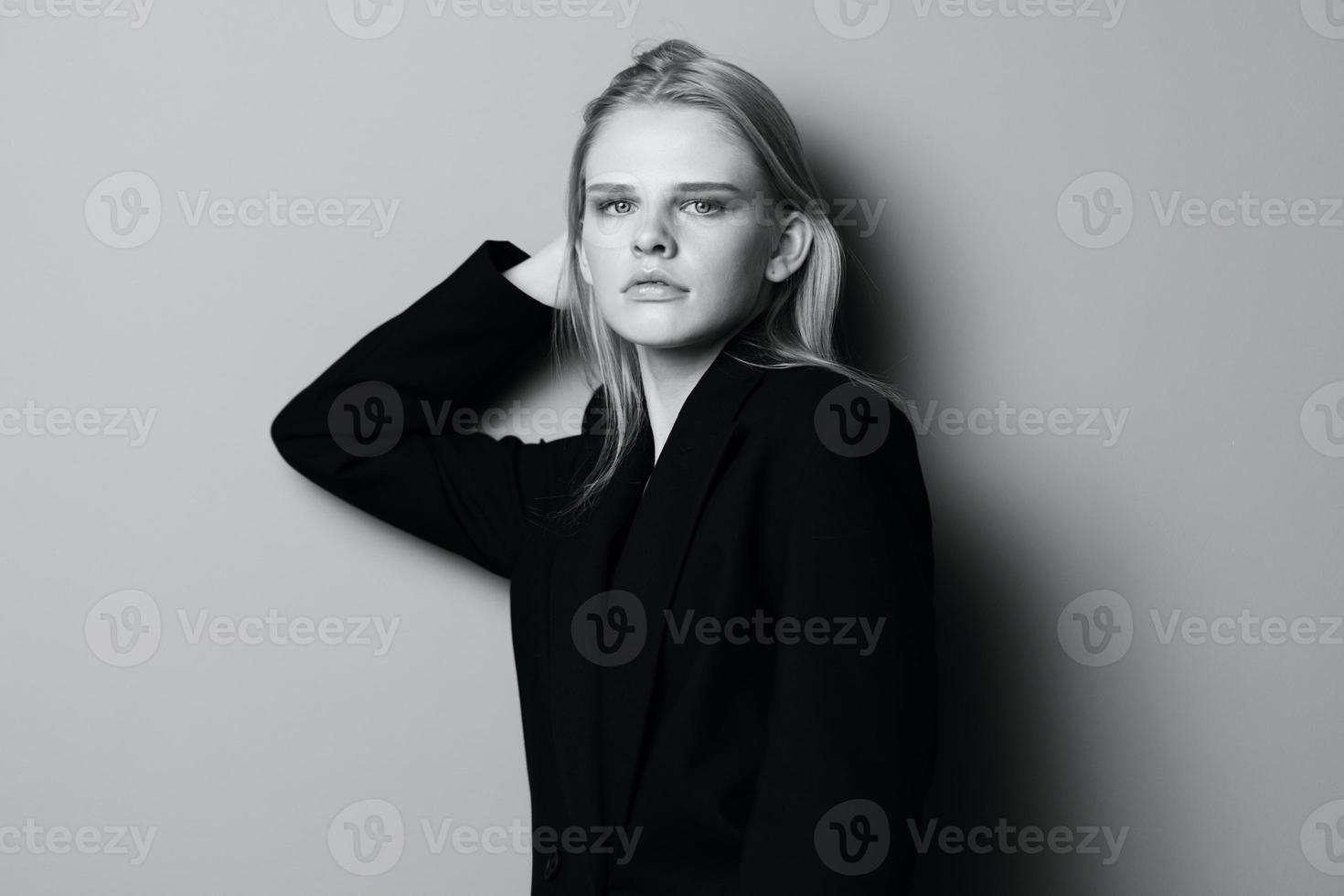 sexy grave joven mujer en negro chaqueta se inclina en el estudio pared conmovedor pelo mira a cámara. elegante concepto para Moda marcas bandera foto