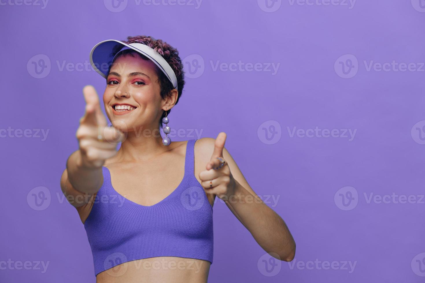 Portrait of a sporty fashion woman posing smiling with teeth and pointing a finger at the camera in a purple yoga tracksuit and a transparent cap on a purple monochrome background photo