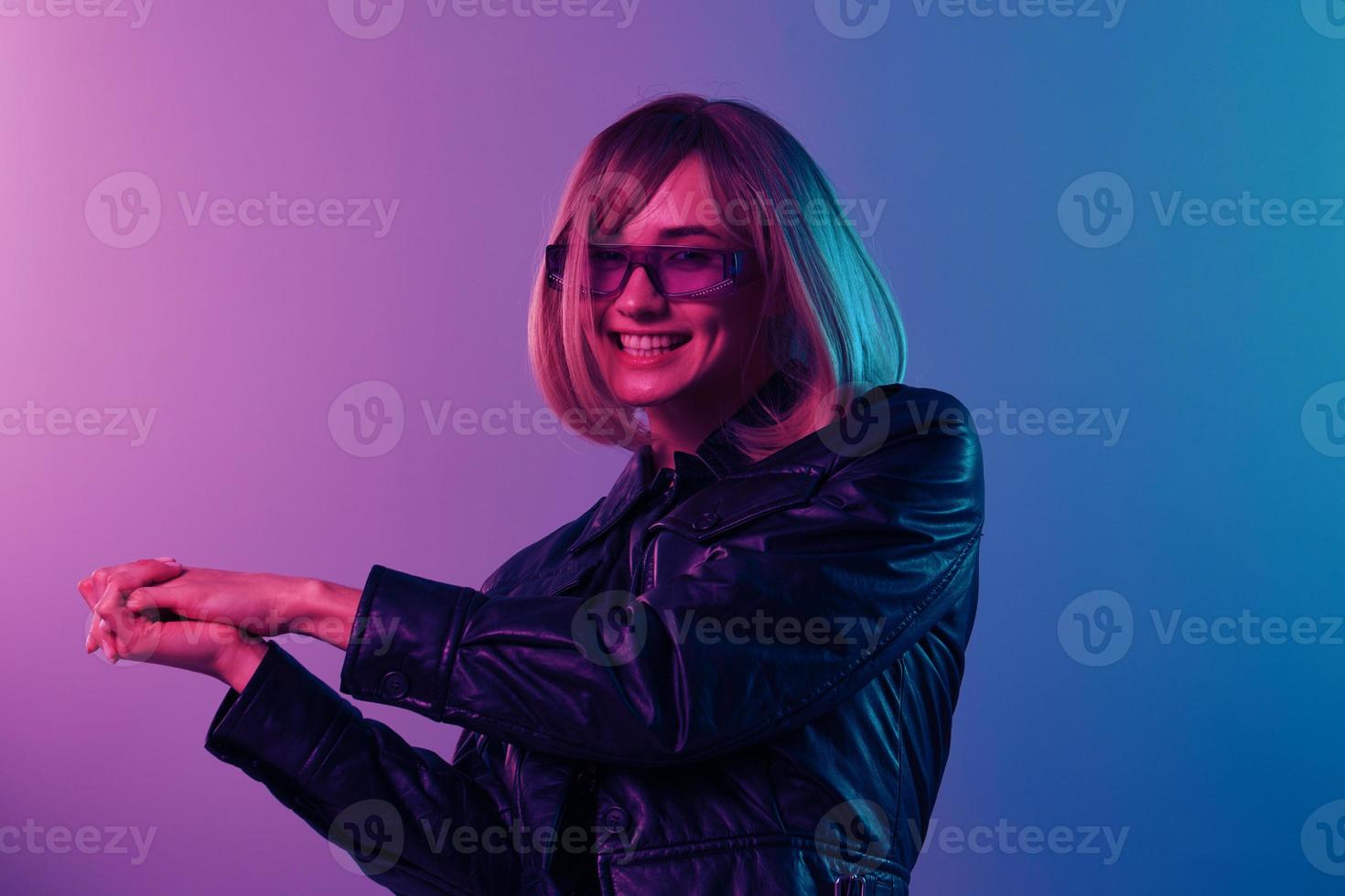 linda hermosa rubia mujer en cuero chaqueta brillante vestir de moda Gafas de sol doblez manos Mira a cámara posando aislado en azul rosado color ligero estudio antecedentes. neón fiesta concepto. Copiar espacio foto