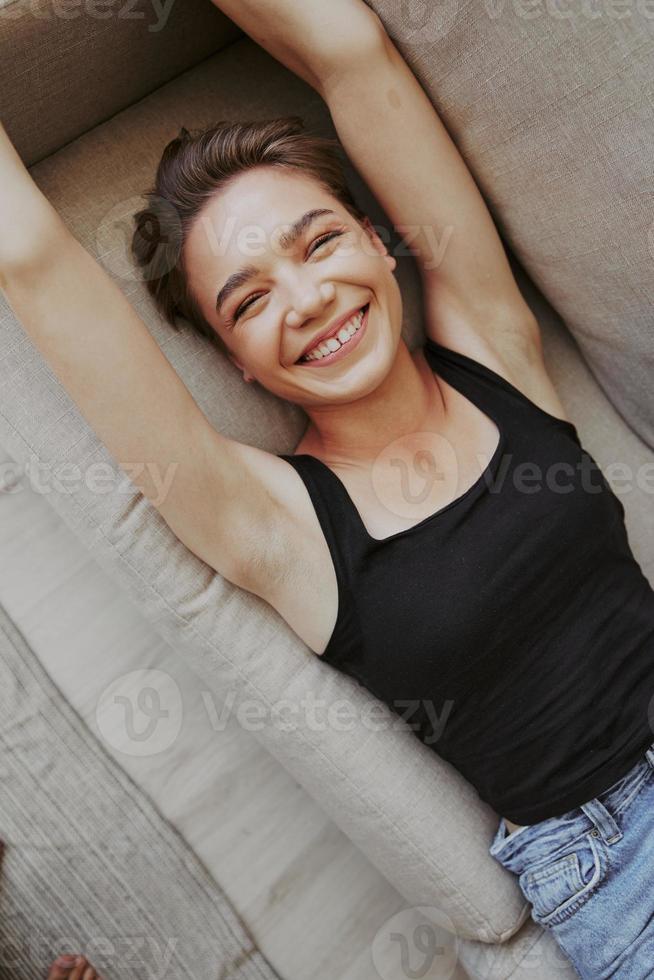 joven mujer con corto Corte de pelo pelo teniendo divertido a hogar en el sofá sonrisa y felicidad, vacaciones a hogar, natural posando sin filtros, gratis Copiar espacio foto