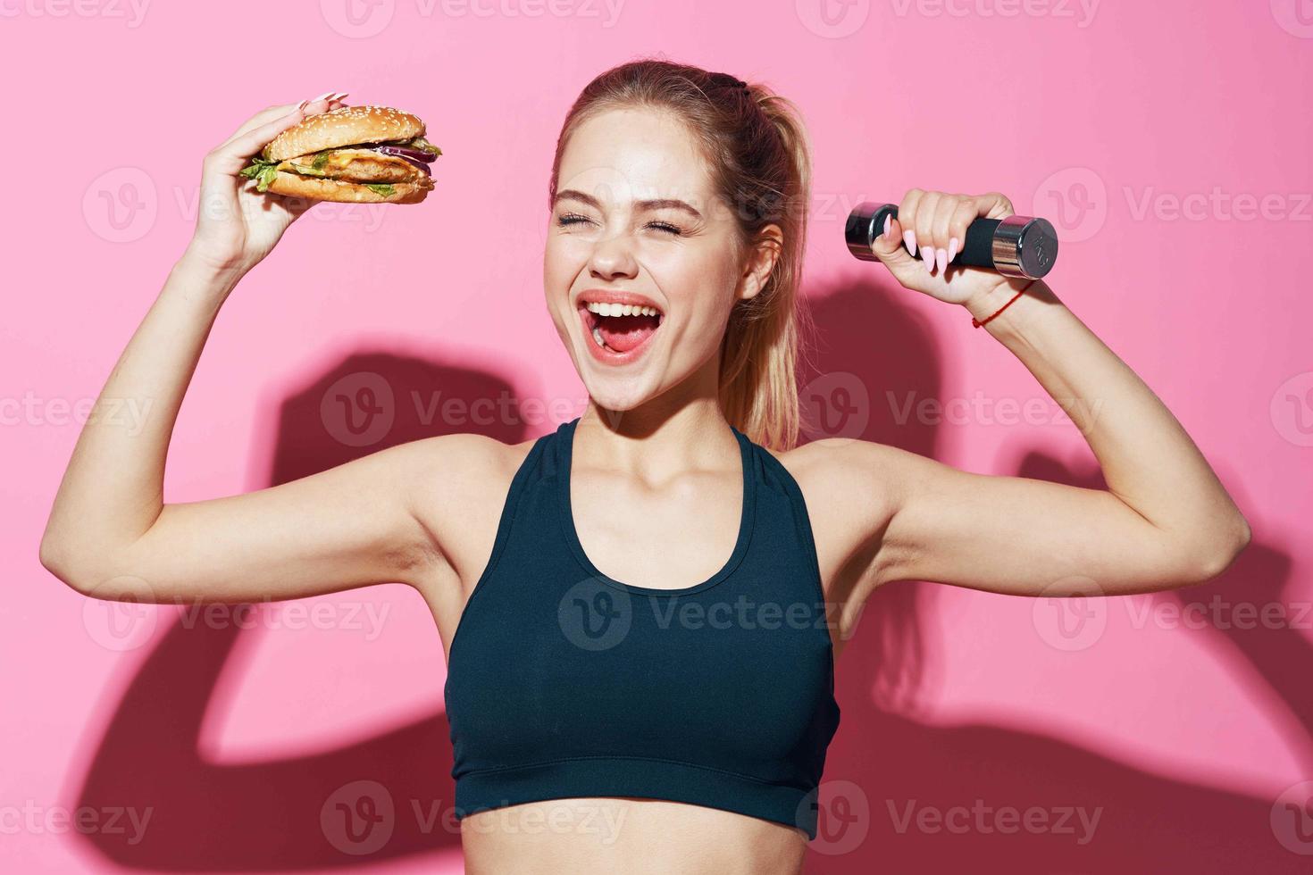 sportive woman with hamburger in hands eating food pink background fast food photo