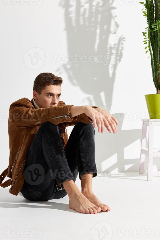 hermoso joven hombre se sienta en el piso en un brillante habitación y un flor en un maceta foto